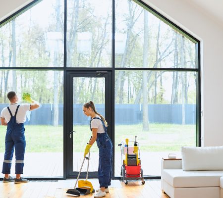 Young cleaners in blue uniform mopping wooden floor, cleaning big panoramic window, preparing country house for selling. Successful cleaning service concept
